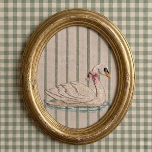 May include: An oval, gold-framed embroidery featuring a white swan with a pink bow around its neck, floating on blue water. The background is striped, and the frame is set against a green and white checkered fabric.