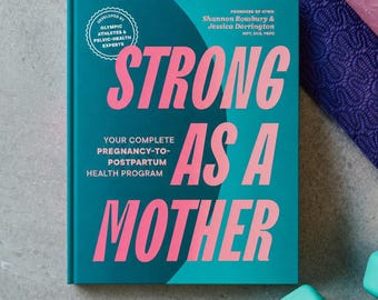 Può includere: Un libro turchese intitolato "STRONG AS A MOTHER" con lettere rosa, che promuove un programma di salute post-partum. Il libro è accanto a un tappetino da yoga viola e due manubri turchesi. La copertina del libro indica che è stato sviluppato da atleti olimpici.