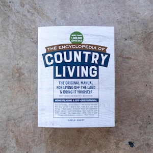 May include: A book titled "The Encyclopedia of Country Living" with the subtitle "The Original Manual for Living Off the Land & Doing It Yourself." The cover is white with blue and brown text. It includes the text "More than 1,000,000 copies sold."
