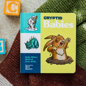 May include: A children's book titled "Cryptid Babies" with illustrations of mythical creatures. The cover is yellow and blue, with the title in large letters. Wooden alphabet blocks with the letters A, B, and C are scattered around the book.