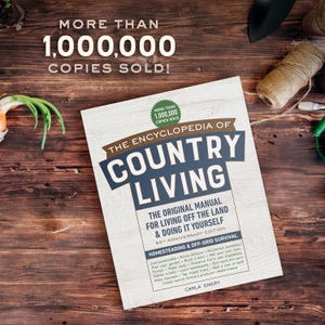 May include: The Encyclopedia of Country Living book, with the text "More than 1,000,000 copies sold!" on a wooden surface. The book's cover features the title in large blue letters. The book is the original manual for living off the land and doing it yourself.