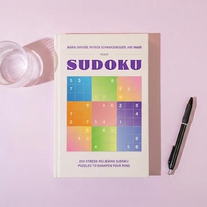 May include: A Sudoku puzzle book with a colorful grid design, a black pen, and a clear glass on a light purple background. The book's cover features the title "SUDOKU" in purple and the text "200 Stress-Relieving Sudoku Puzzles to Sharpen Your Mind."