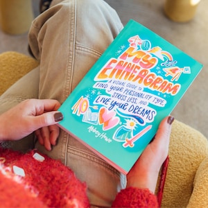 May include: A teal book titled "My Enneagram" with colorful illustrations and the text "A Visual Guide to Find Your Personality Type, Stress Less, and Live Your Dreams." The book is held in a person's hands.