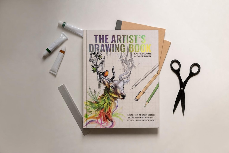 May include: An open copy of "The Artist's Drawing Book" with a deer illustration on the cover, surrounded by art supplies including paint tubes, pencils, scissors, and a notepad. The book's cover features the text "The Artist's Drawing Book".