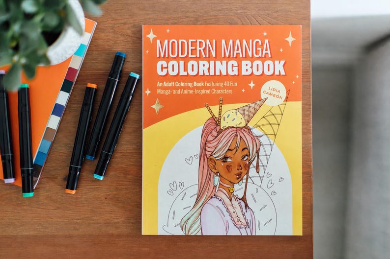May include: A Modern Manga Coloring Book with an illustration of a manga-style character with ice cream on their head. The book is on a wooden surface with colorful markers and a notebook.