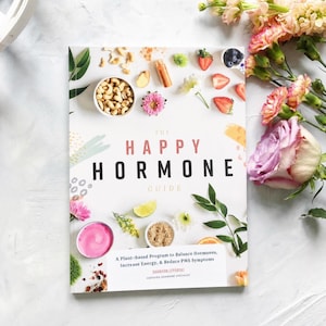 May include: A book titled "The Happy Hormone Guide" with text that reads "A Plant-based Program to Balance Hormones, Increase Energy, & Reduce PMS Symptoms." The book is surrounded by flowers, nuts, and fruit.