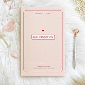May include: A pale pink journal with the words "don't cramp my style" and a small red heart. The journal is part of "the hoppy hormone tracker" series, a wellness journal for monthly cycle tracking. A silver pen with a diamond-shaped top rests beside it.
