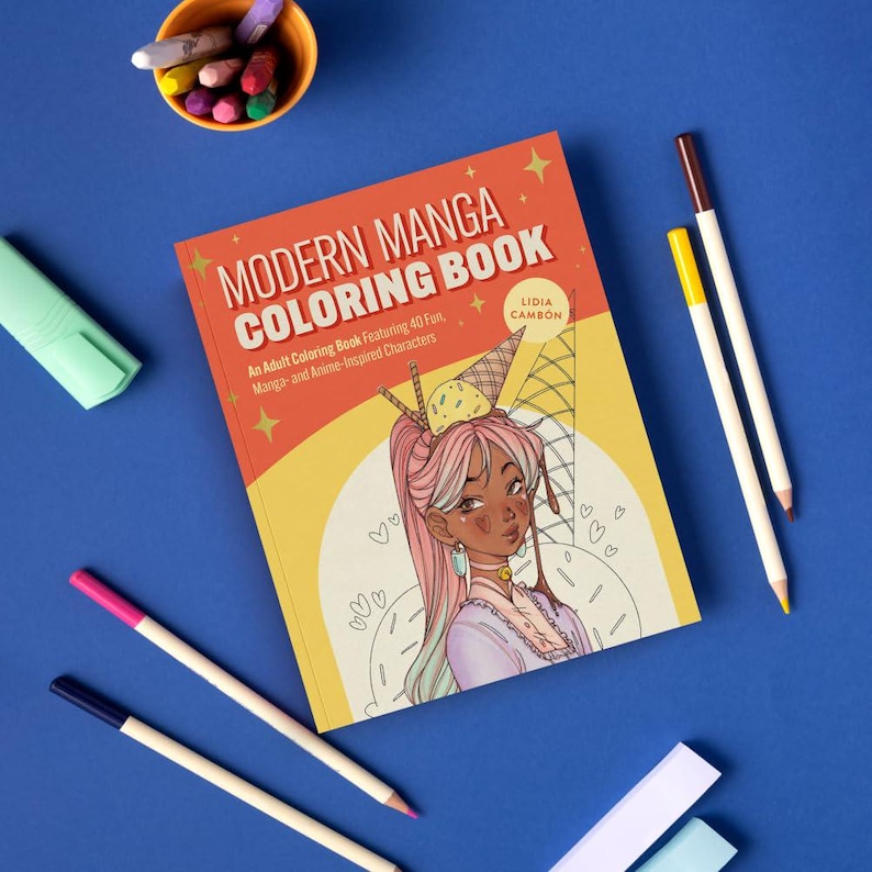May include: A "Modern Manga Coloring Book" with a colorful illustration of a manga-style character with pink hair and an ice cream cone on her head. The book is on a blue surface with colored pencils and a highlighter.