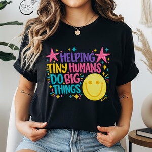 Può includere: Una t-shirt nera con una stampa grafica colorata con il testo "HELPING TINY HUMANS DO BIG THINGS" in viola, giallo, rosa e blu, accentuata da stelle rosa e una grande icona di faccina sorridente gialla con scintille blu e gialle.