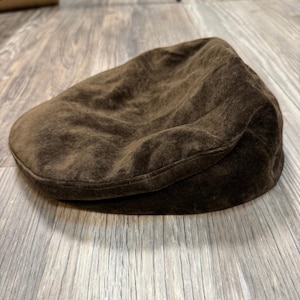 Puede incluir: Una gorra plana de ante marrón oscuro con una corona redondeada y una visera corta y rígida. La superficie exterior tiene un tacto suave y texturizado característico del ante, con costuras visibles a lo largo de los bordes de la visera.