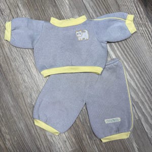 May include: A light gray two-piece outfit consisting of a long-sleeved pullover top and matching pants. The top features a yellow collar, yellow sleeve cuffs, and a small embroidered white cat graphic. The pants have yellow ankle cuffs.