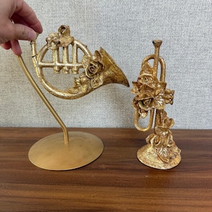 May include: Two gold-colored decorative musical instruments. One is a French horn on a stand, the other a trumpet. Both are adorned with rose details. The instruments are displayed on a wooden surface.