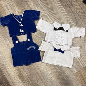 Op de afbeelding: Een set poppenkleding bestaande uit een koningsblauw fluwelen vestje met witte bies en twee knopen, een bijpassende blauwe fluwelen tuinbroek met Cabbage Patch Kids-borduursel, en twee witte shirts met korte mouwen, kragen, ruches en vlinderdassen.