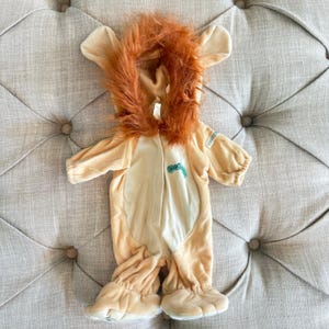 May include: A light tan plush lion costume featuring a hood with a thick, shaggy orange faux fur mane. The one-piece garment has a cream-colored chest panel, long sleeves, and attached foot covers. A small teal embroidered logo is on the chest.
