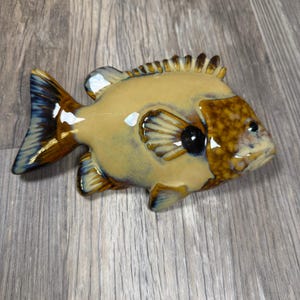 May include: A ceramic fish figurine featuring a glossy finish with a cream-colored body, a brown mottled head, and dark blue-tipped fins. The fish has a prominent dorsal fin, a rounded tail, and a dark eye, showcasing a smooth, reflective surface.