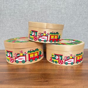 Puede incluir: Tres cajas de Navidad de madera con tapas. Cada caja está decorada con un diseño de tren colorido con Papá Noel, bastones de caramelo y la palabra "NOEL". Las cajas son de forma ovalada y se encuentran sobre una superficie de madera.