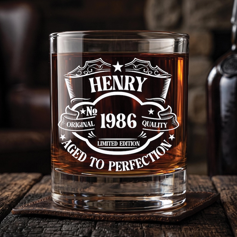 May include: A clear glass tumbler filled with amber liquid, featuring white etched text that reads HENRY, No 1986, ORIGINAL QUALITY, LIMITED EDITION, and AGED TO PERFECTION, arranged within a decorative frame with stars and scrollwork elements.