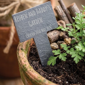 Puede incluir: Un marcador de jardín de pizarra gris oscuro con el texto "REUBEN AND DADDY'S GARDEN" y "CREATED 20TH MAY 2026" se encuentra en una maceta de terracota con tierra y plantas verdes. El marcador tiene forma de T.