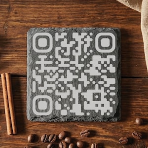 Könnte beinhalten: Ein quadratischer, dunkelgrauer Schieferuntersetzer mit einem QR-Code-Design in Hellgrau. Der Untersetzer ist von Kaffeebohnen und Zimtstangen auf einer Holzoberfläche umgeben. Der Untersetzer ist etwa 10 cm groß.