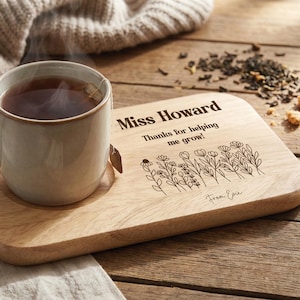 Può includere: Una tazza in ceramica con tè e una bustina di tè è appoggiata su un vassoio di legno. Il vassoio è inciso con il nome "Miss Howard", il messaggio "Thanks for helping me grow!" e un motivo floreale. Il vassoio è firmato "From Evie".