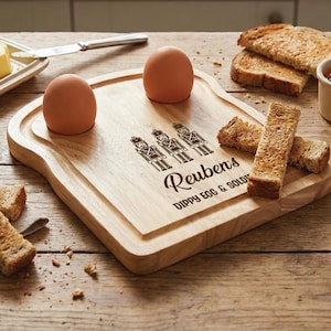 May include: A light-coloured wooden egg and toast board shaped like a slice of bread with two circular indentations for eggs. The board features a laser-engraved design of three nutcracker soldiers and the text "Reuben's DIPPY EGG & SOLDIERS".
