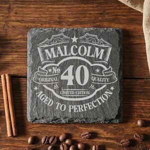 May include: A square slate coaster with a rough, natural edge featuring an engraved design. The text reads MALCOLM, No 40, ORIGINAL QUALITY, LIMITED EDITION, and AGED TO PERFECTION, arranged in a decorative label style with stars and flourishes.