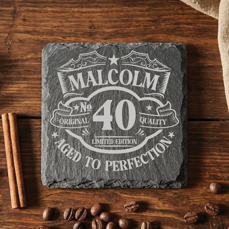 May include: A square slate coaster with a rough, natural edge featuring an engraved design. The text reads MALCOLM, No 40, ORIGINAL QUALITY, LIMITED EDITION, and AGED TO PERFECTION, arranged in a decorative label style with stars and flourishes.