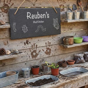 May include: A wooden mud kitchen with a slate sign reading "Reuben's Mud Kitchen", featuring insect drawings. The kitchen has shelves with metal buckets, gardening tools, bowls, and planting supplies. Handprints are visible on the wooden surface, creating a playful aesthetic.