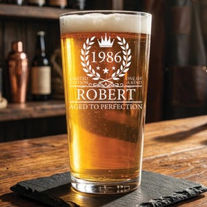 May include: A clear glass pint beer mug filled with golden beer, featuring white etched text that reads 1986, LIMITED EDITION, ONE OF A KIND, ROBERT, and AGED TO PERFECTION, along with a crown and laurel wreath graphic design.