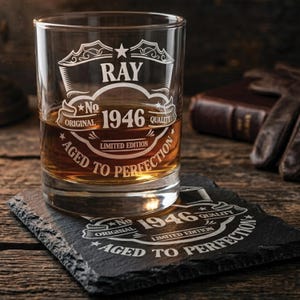May include: A clear glass tumbler filled with amber liquid, featuring white etched text reading RAY, No 1946, ORIGINAL, QUALITY, LIMITED EDITION, and AGED TO PERFECTION. It sits on a matching black slate coaster with the same engraved text design.