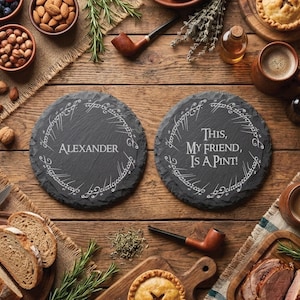 Op de afbeelding: Twee ronde, donkergrijze leistenen onderzetters met witte letters en een decoratieve rand. Eén onderzetter leest "ALEXANDER", de andere zegt "THIS, MY FRIEND, IS A PINT!". De onderzetters zijn omringd door eten en drinken op een houten tafel.