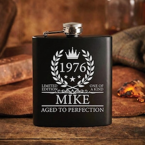 May include: A matte black metal flask featuring a silver engraved design of a laurel wreath, a crown, and the year 1976. The text reads: LIMITED EDITION, ONE OF A KIND, MIKE, and AGED TO PERFECTION. It has a silver screw-top cap with a hinged arm.