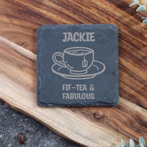 May include: A dark grey slate coaster with the name "JACKIE" and a tea cup design etched on it. The text "FIF-TEA & FABULOUS" is below the design. The coaster is displayed on a wooden surface.