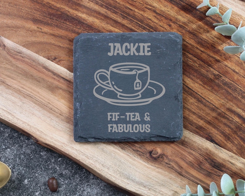 May include: A dark grey slate coaster with the name "JACKIE" and a tea cup design etched on it. The text "FIF-TEA & FABULOUS" is below the design. The coaster is displayed on a wooden surface.
