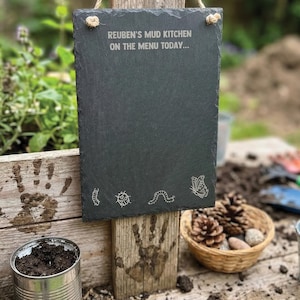 Może przedstawiać: Prostokątna tabliczka z ciemnoszarego łupka o nierównych krawędziach, z wygrawerowanym napisem "REUBEN'S MUD KITCHEN ON THE MENU TODAY..." oraz małymi rysunkami liniowymi gąsienicy, biedronki, robaka i motyla u dołu.