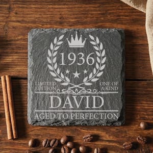 May include: A square, dark gray slate coaster with white engraved text and design. The design includes the year 1936, a crown, laurel wreath, stars, and the name "DAVID". The text reads "LIMITED EDITION" and "AGED TO PERFECTION".