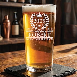 May include: A clear glass pint beer mug filled with golden beer and white foam. The glass features white etched text reading 2005, LIMITED EDITION, ONE OF A KIND, ROBERT, and AGED TO PERFECTION, along with a crown and laurel wreath graphic design.