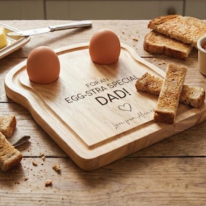 Puede incluir: Una tabla de desayuno de madera con forma de rebanada de pan, con dos huevos marrones y tostadas. La tabla tiene el texto "FOR AN EGG-STRA SPECIAL DAD!" y un símbolo de corazón. También están grabadas las palabras "Love from Alice xx".