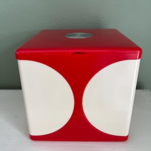 Peut inclure: Un conteneur cubique rouge et blanc avec un accent circulaire argenté sur le dessus. Le conteneur a un couvercle rouge et des côtés blancs avec deux grandes formes blanches incurvées. Le design est moderne et géométrique.