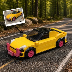 Op de afbeelding: Een gele, van bouwstenen gemaakte modelauto met een zwart dak, roze wielen en een roze voorbumper. Een kleine foto van een echte gele auto met roze wielen is met een zwarte pijl aan het model bevestigd.