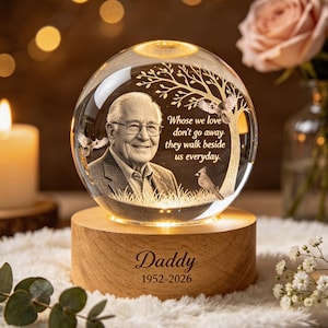 May include: A spherical glass ornament featuring an etched portrait of an older man, a tree, and two birds. The text reads, "Whose we love don't go away, they walk beside us everyday." The base is light wood engraved with "Daddy" and "1952-2026."