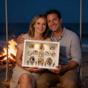 May include: A couple holds a white framed shadow box with miniature framed photos and figurines. The box has the text "Love you forever, happy anniversary." The background includes a beach, ocean, and a bonfire.
