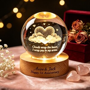 May include: A clear glass sphere resting on a circular wooden base, featuring an internal laser-etched heart surrounded by clouds and the text: Clouds wrap the heart, I wrap you in my arms. The base is engraved with: Avire & Jack Happy 1st Anniversary.