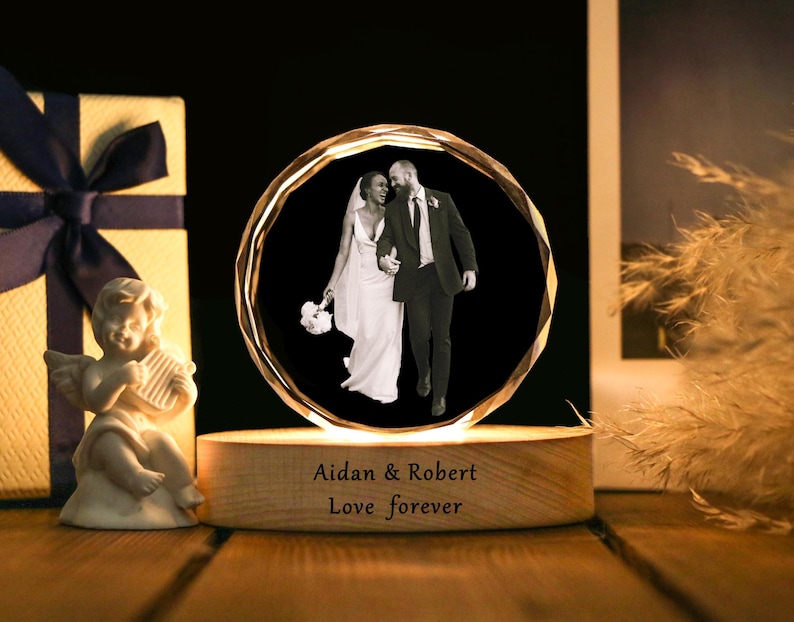 May include: A crystal sphere with a black and white photo of a couple in wedding attire. The sphere sits on a wooden base with the engraved text "Aidan & Robert Love forever". A gift box with a dark blue ribbon and a small angel figurine are in the background.