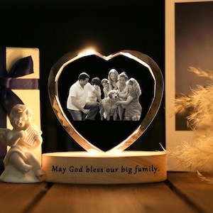 May include: A heart-shaped crystal photo frame with a family portrait inside, illuminated by a warm light. The wooden base has the inscription "May God bless our big family." A gift box and a small angel figurine are also present.