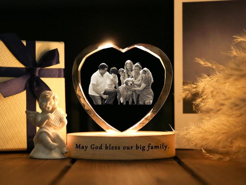 May include: A heart-shaped crystal photo frame with a family portrait inside, illuminated by a warm light. The wooden base has the inscription "May God bless our big family." A gift box and a small angel figurine are also present.