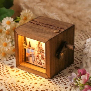 Peut inclure: Boîte à musique en bois avec une scène de Noël éclairée à l'intérieur. La boîte est en forme de cube avec une manivelle sur le côté et du texte sur le dessus. Une petite figurine d'ange blanc et des fleurs sont à proximité.