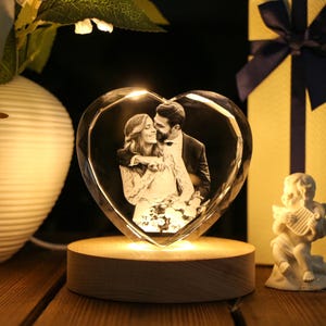 May include: A heart-shaped crystal photo frame with a couple's image, illuminated by a warm light. The frame sits on a wooden base, with a small cherub figurine and a wrapped gift box in the background. The overall theme is romance and love.