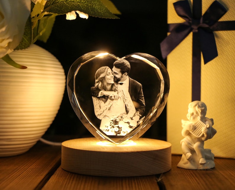 May include: A heart-shaped crystal photo frame with a couple's image, illuminated by a warm light. The frame sits on a wooden base, with a small cherub figurine and a wrapped gift box in the background. The overall theme is romance and love.