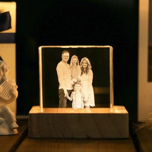 May include: A family portrait is displayed within a clear, rectangular crystal block, illuminated from below by a wooden base. The image shows a family of four. A small, white cherub statue with a harp is on the left, and a gift box is in the background.